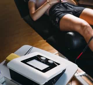 Patientin erhält Physikalische Therapie mit Elektroden am Knie, Therapiegerät steht daneben.