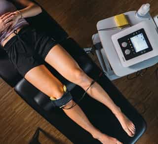 Patientin liegt auf Liege bei PhysioSport, Elektroden am Knie für Elektrotherapie in der Physikalischen Therapie, Gerät daneben.