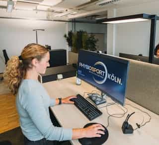 Zu sehen ist der Einblick in einen offenen Arbeitsplatz bei Physiosport Köln. Dort sitzt eine Mitarbeiterin konzentriert am Computer. Im Hintergrund eine weitere Mitarbeiterin.