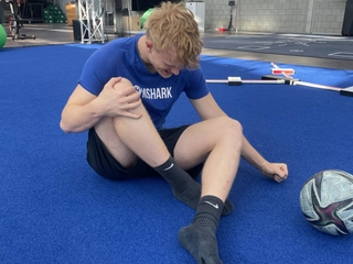 Man sieht einen Sportler bei PhysioSport Köln, schräg von vorne gesehen, der sich vor Schmerzen an sein Knie fasst und mit einem Arm am Boden abstützt..