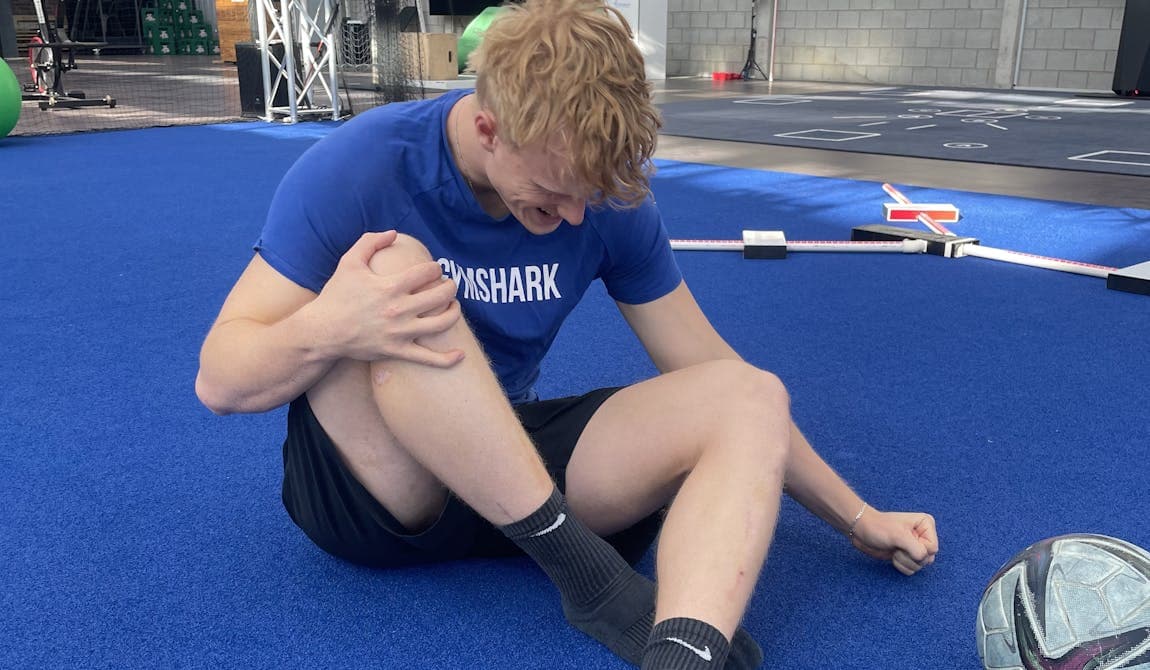 Man sieht einen Sportler bei PhysioSport Köln, schräg von vorne gesehen, der sich vor Schmerzen an sein Knie fasst und mit einem Arm am Boden abstützt..