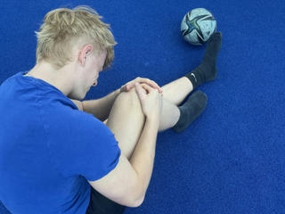 Man sieht einen Sportler bei PhysioSport Köln, schräg von hinten gesehen, der sich vor Schmerzen an sein Knie fasst.